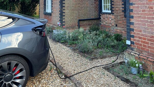 A grey electric car charging on a gravel driveway outside a brick house, with the cable running from the car to a small charge point on the wall