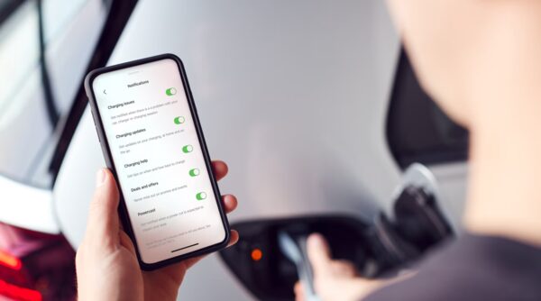 A person holding a smartphone showing an EV charging app notifications screen while plugging a cable into an electric car