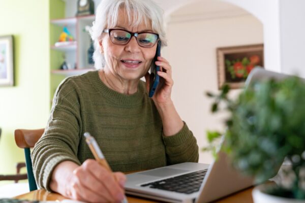 An older woman with glasses talking on a mobile phone while taking notes at a laptop at home