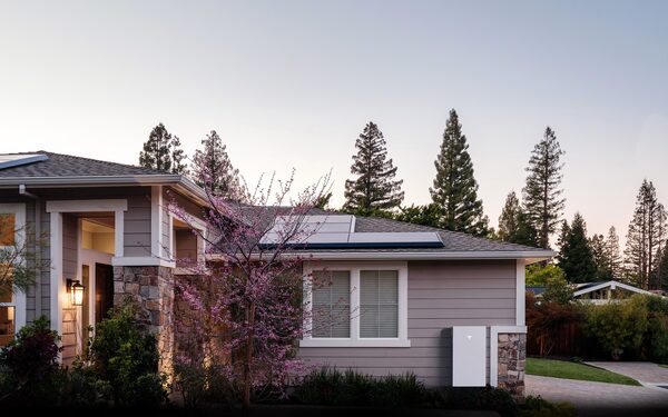 A Tesla Powerwall battery mounted on the exterior wall of a house with solar panels on the roof at dusk