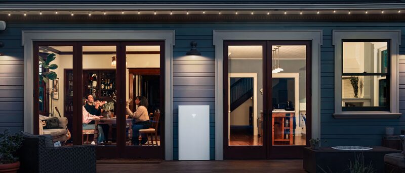 A white Tesla Powerwall home battery mounted on the exterior wall of a house at night, with people visible through the lit windows